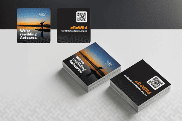 Two stacks of promotional cards on a textured surface. One stack features an image of a silhouette of a person fishing with the text "We're rewilding Aotearoa," and the other with #ReWild and a QR code.