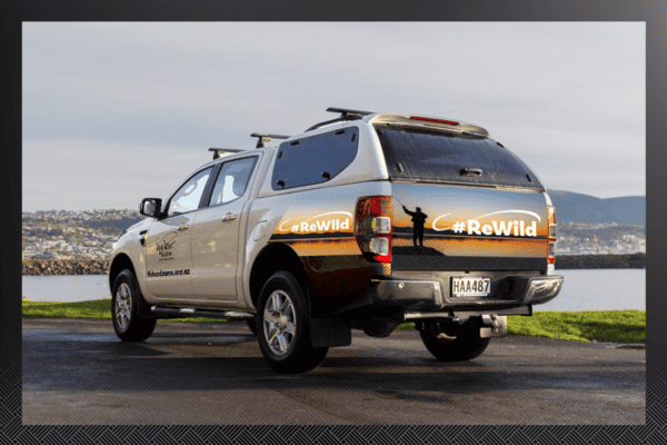 A white utility vehicle with "#ReWild" decals is parked by a body of water with a cityscape in the background. The vehicle has a canopy and a New Zealand license plate reading "HAA487".