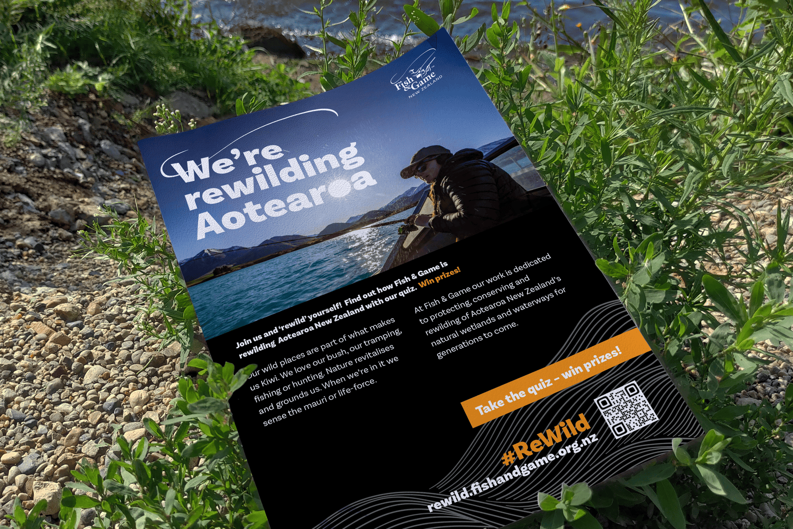 A flyer titled "We're rewilding Aotearoa" lies on some greenery near a body of water. It features an image of a person in nature with mountains and water in the background. The flyer promotes conservation efforts and includes a QR code for more information.